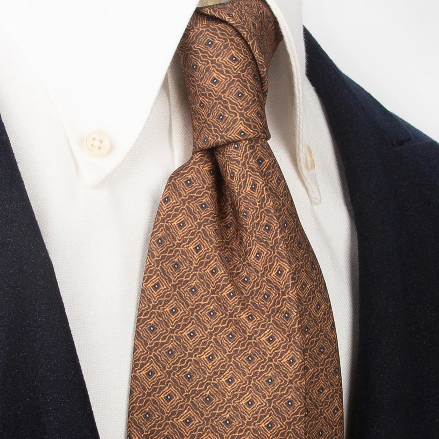 A neatly rolled brown men's necktie with an intricate repeating geometric pattern in gold and dark blue is displayed against a plain white background.