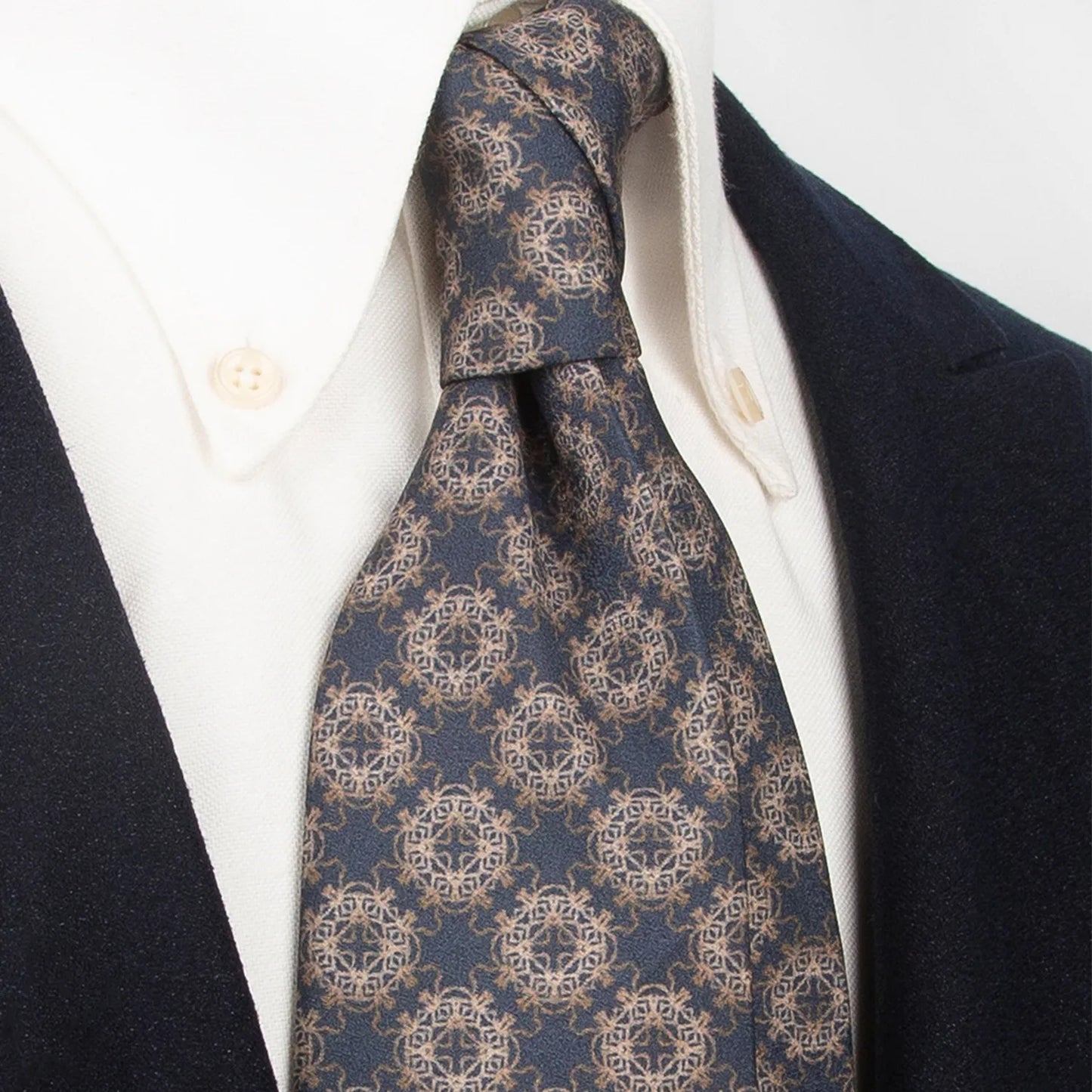 A deep blue men's necktie showcasing its refined golden and brown totem geometric design is tied in a Windsor knot on a crisp white button-down shirt under a dark navy blazer.