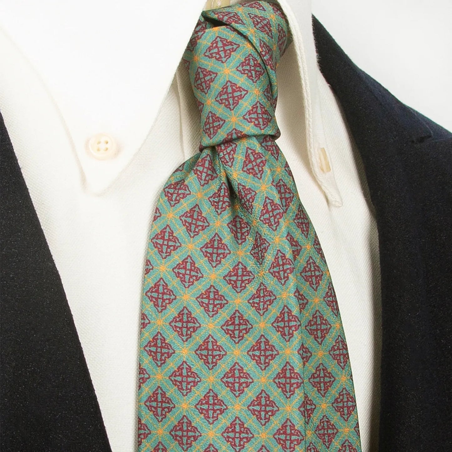 A sophisticated green men's necktie from XueDouLing Tie with an intricate geometric design in red and gold is tied in a four-in-hand knot on a crisp white button-down shirt under a dark navy blazer, suitable for business and special occasions.