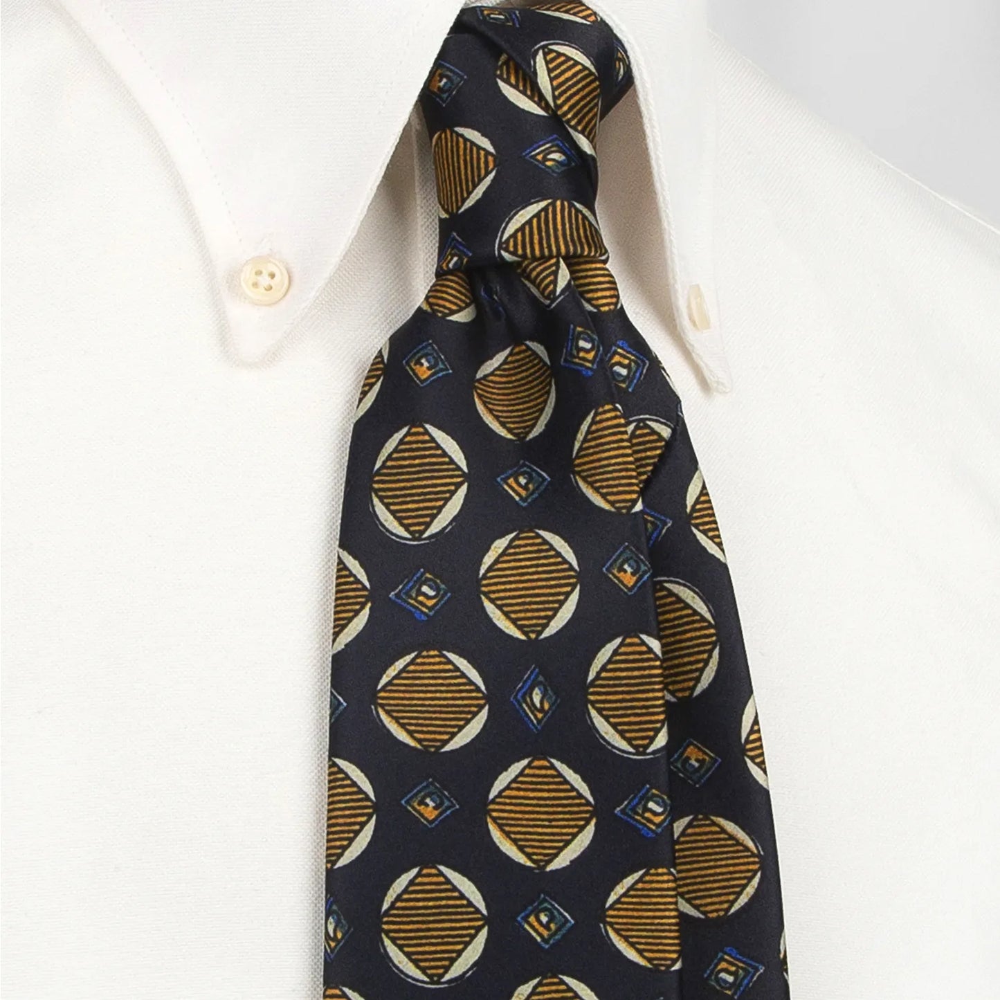 A dark blue men's necktie featuring a modern geometric pattern with circles and diamonds in gold, beige, and blue is tied in a four-in-hand knot on a crisp white button-down shirt, suitable for business and stylish events.