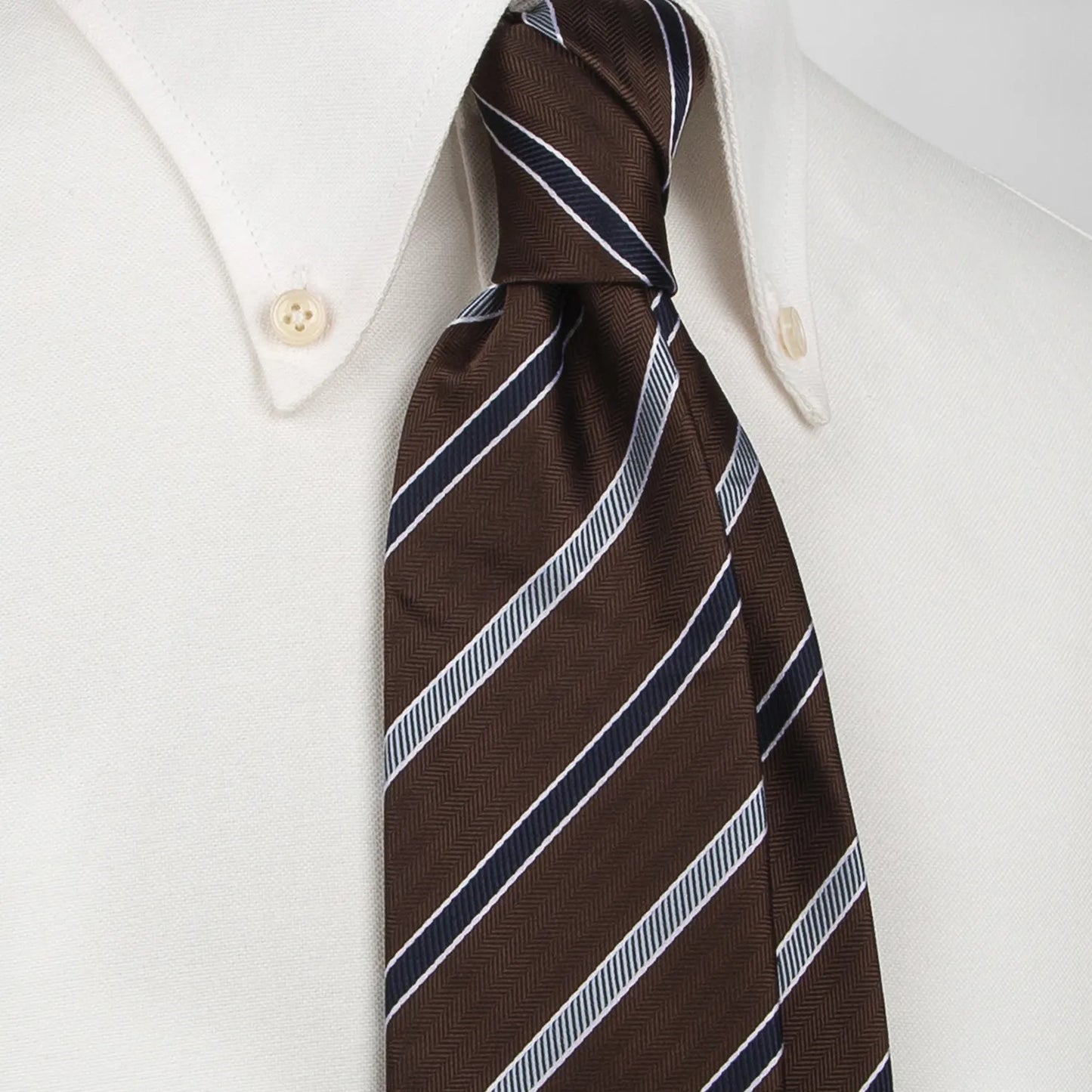 A close-up shows a brown men's necktie with textured diagonal stripes in white and navy blue tied in a four-in-hand knot on a crisp white button-down shirt, suitable for business and professional settings.