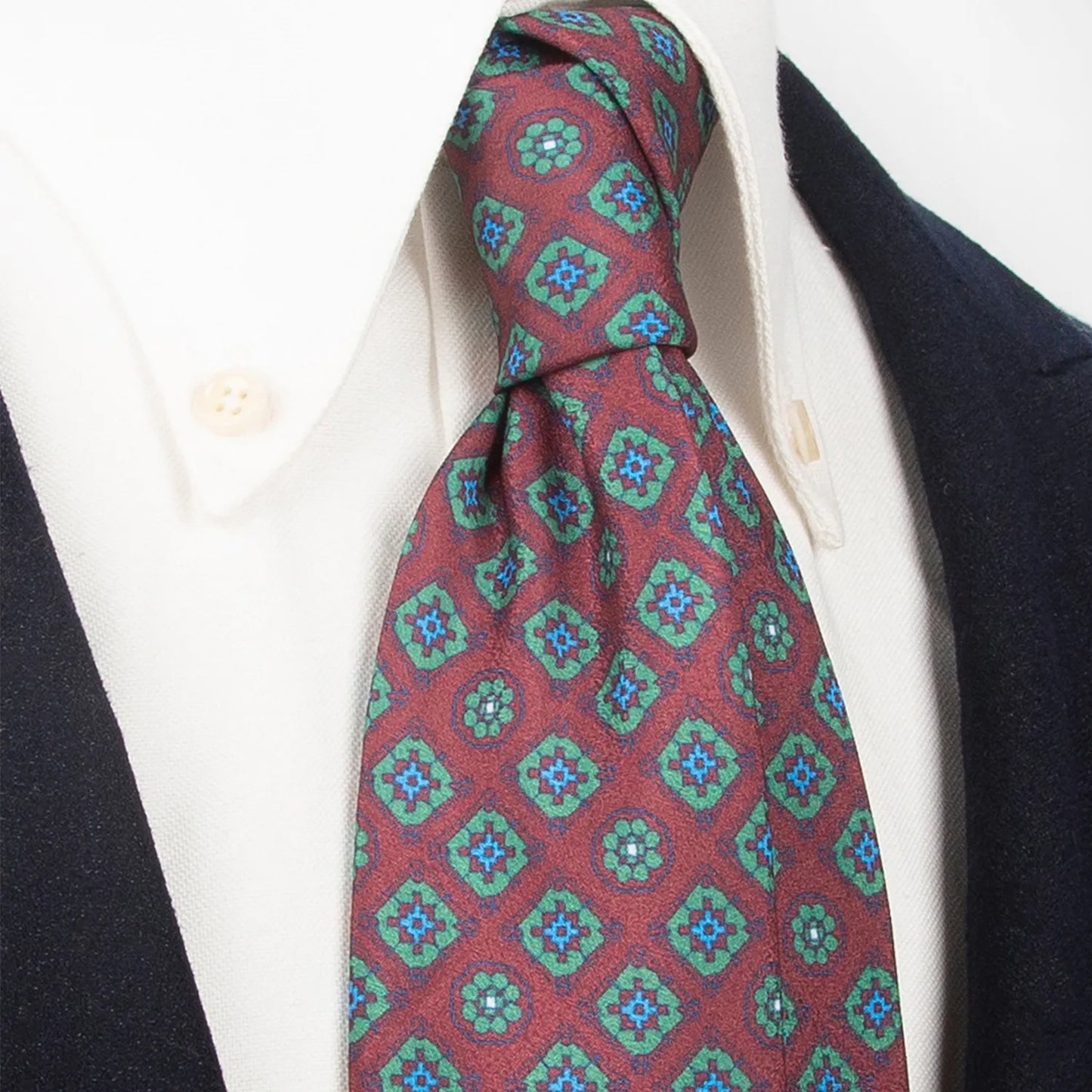 A refined burgundy men's necktie from XueDouLing Tie with an intricate geometric floral design in green and blue is tied in a four-in-hand knot on a crisp white button-down shirt under a dark suit jacket, suitable for business, weddings, and elegant events.