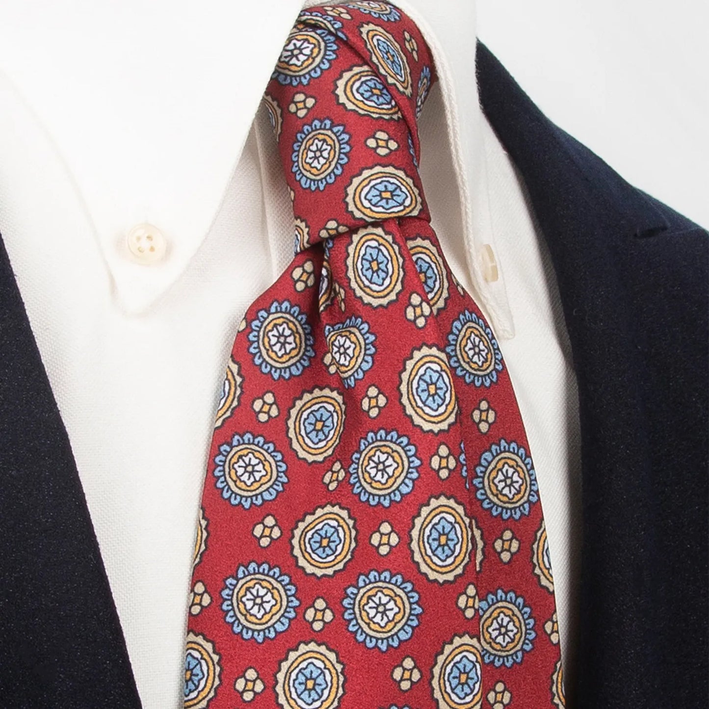 An elegant deep red men's necktie from XueDouLing Tie with a lively pattern of circular floral medallions in blue, yellow, and white, tied in a four-in-hand knot on a crisp white button-down shirt under a dark suit jacket, perfect for business, weddings, and festive events.