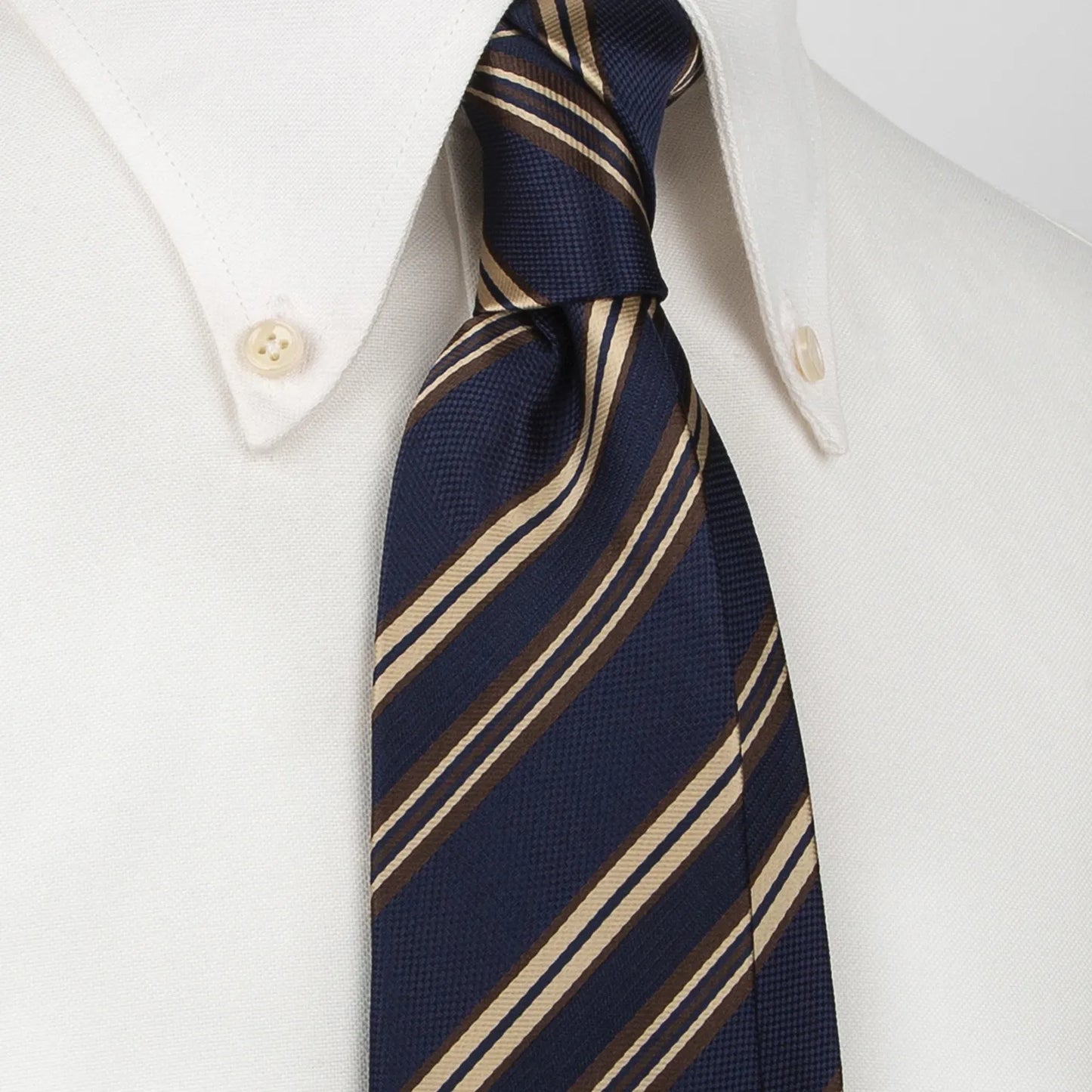 A dark blue men's necktie featuring diagonal stripes in tan and brown, both with a subtle woven texture, is tied in a four-in-hand knot on a crisp white button-down shirt, suitable for business and professional settings.