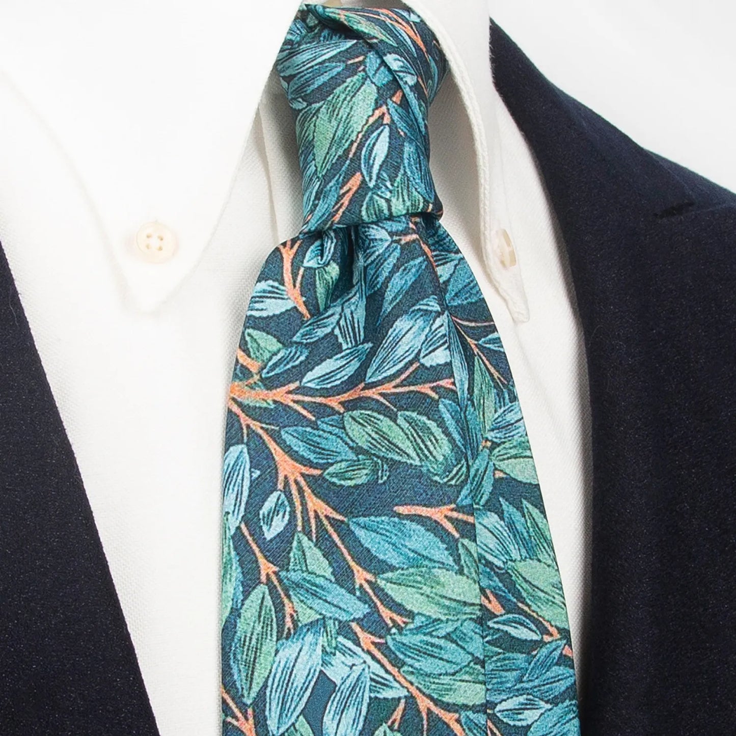 A dark blue men's necktie from XueDouLing Tie showcasing its nature-inspired leaf pattern in green, blue, and brown, tied in a four-in-hand knot on a crisp white button-down shirt under a dark blazer, suitable for business casual and stylish events.