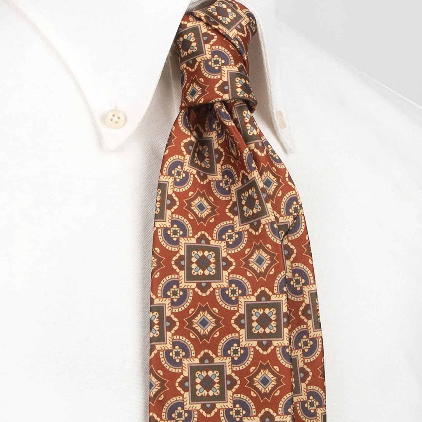 A red-brown men's necktie featuring a complex geometric pattern in blue, beige, and green, incorporating squares, circles, and star-like shapes, is tied in a four-in-hand knot on a crisp white button-down shirt, suitable for business and elegant events.