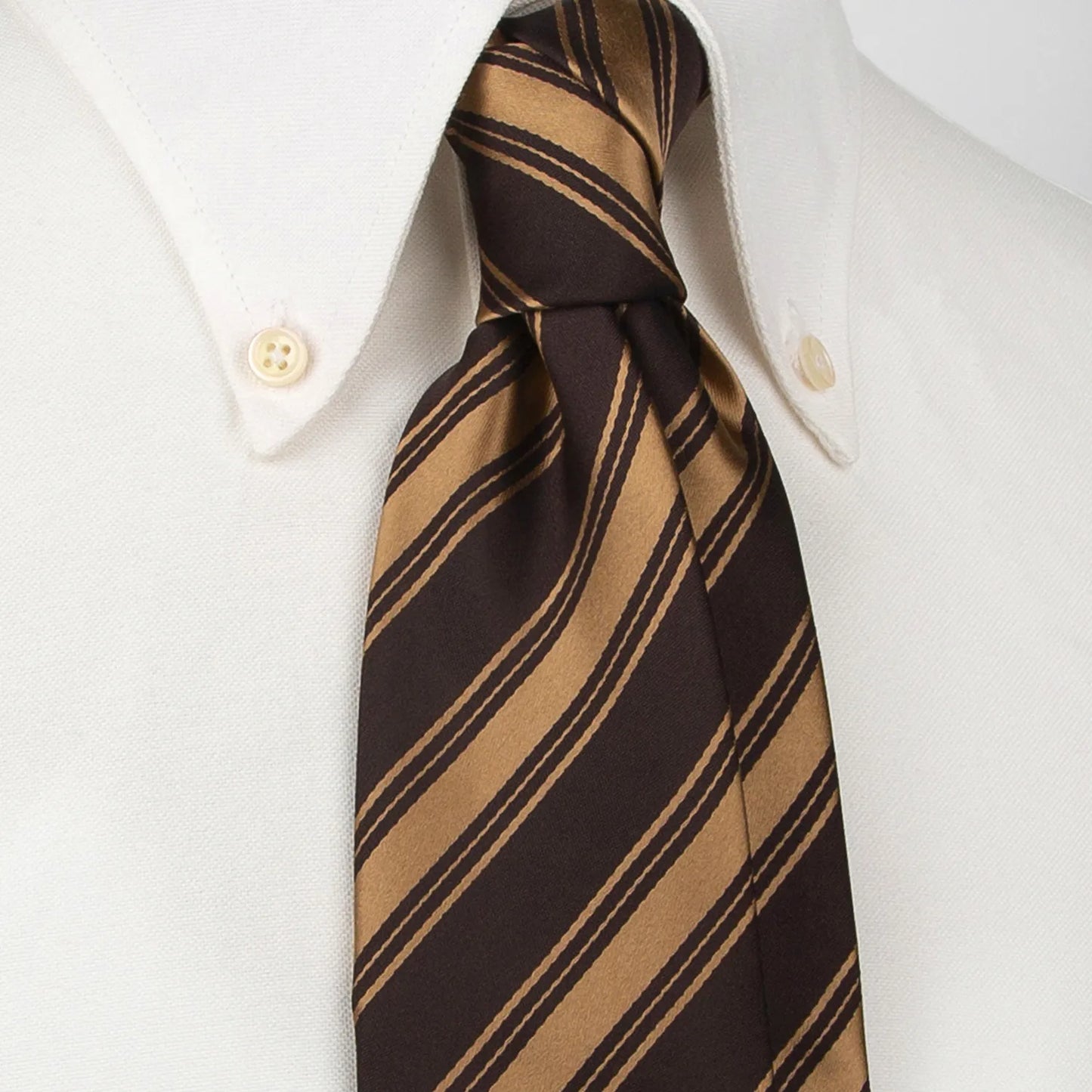 A dark brown men's necktie featuring bold diagonal stripes in a lustrous gold hue is tied in a four-in-hand knot on a crisp white button-down shirt, suitable for business and formal settings.
