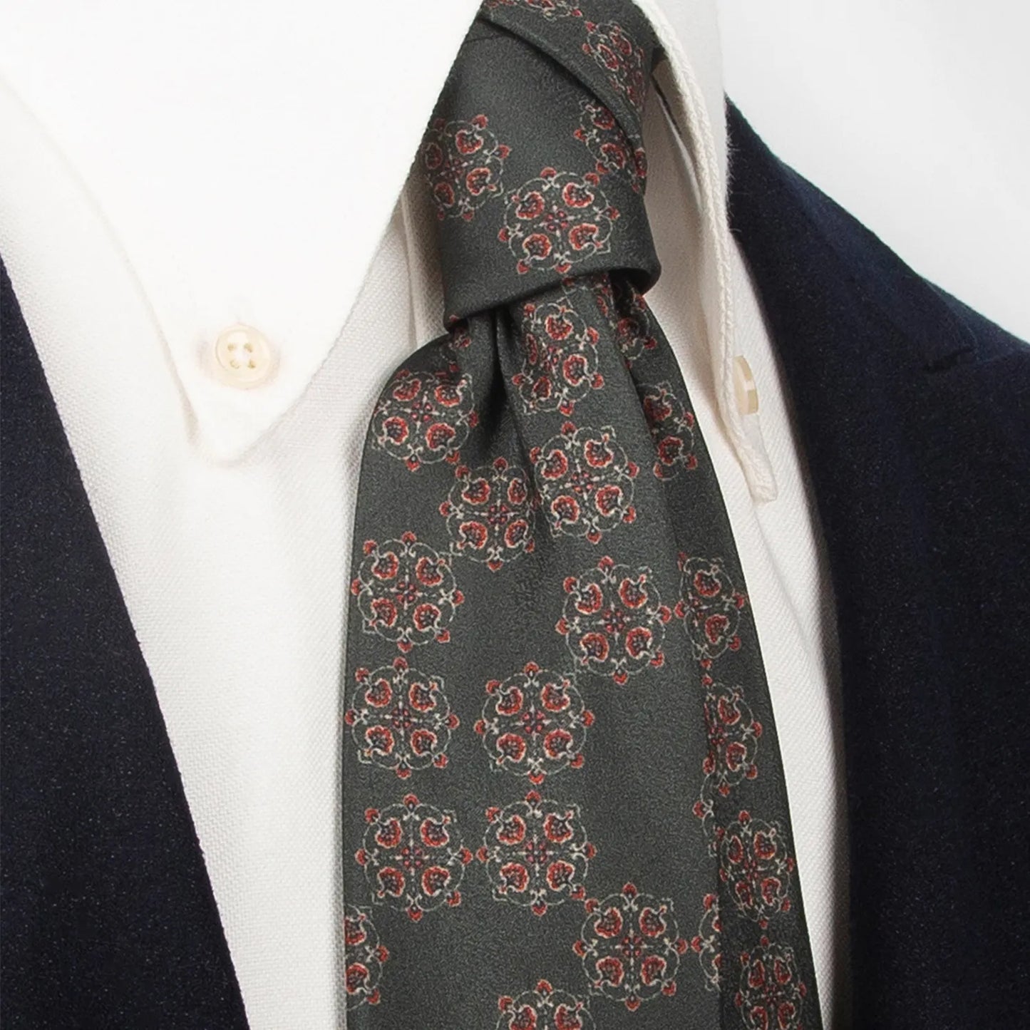 A sophisticated dark grey-green men's necktie from XueDouLing Tie with an intricate retro floral design in red, beige, and brown is tied in a four-in-hand knot on a crisp white button-down shirt under a dark blazer, suitable for business and refined events.