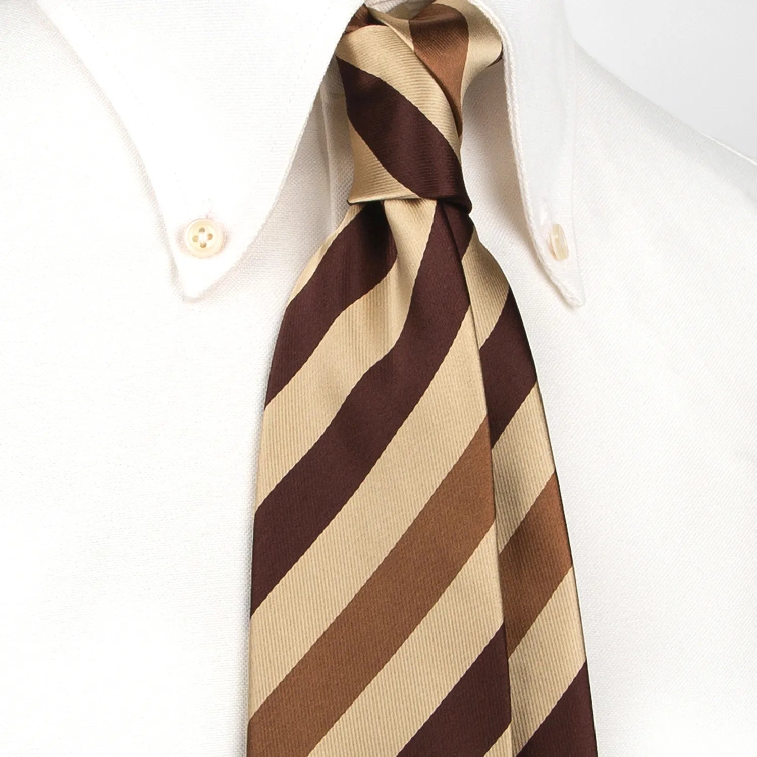 A classic men's necktie with wide diagonal stripes in beige, brown, and dark brown is tied in a four-in-hand knot on a crisp white button-down shirt, suitable for business and professional settings.