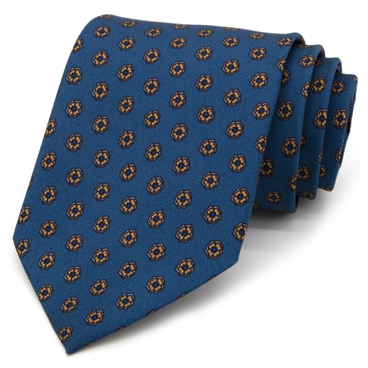 A blue men's necktie featuring a repeating pattern of small brown and beige floral dots is tied in a four-in-hand knot on a crisp white button-down shirt, suitable for business and professional settings.