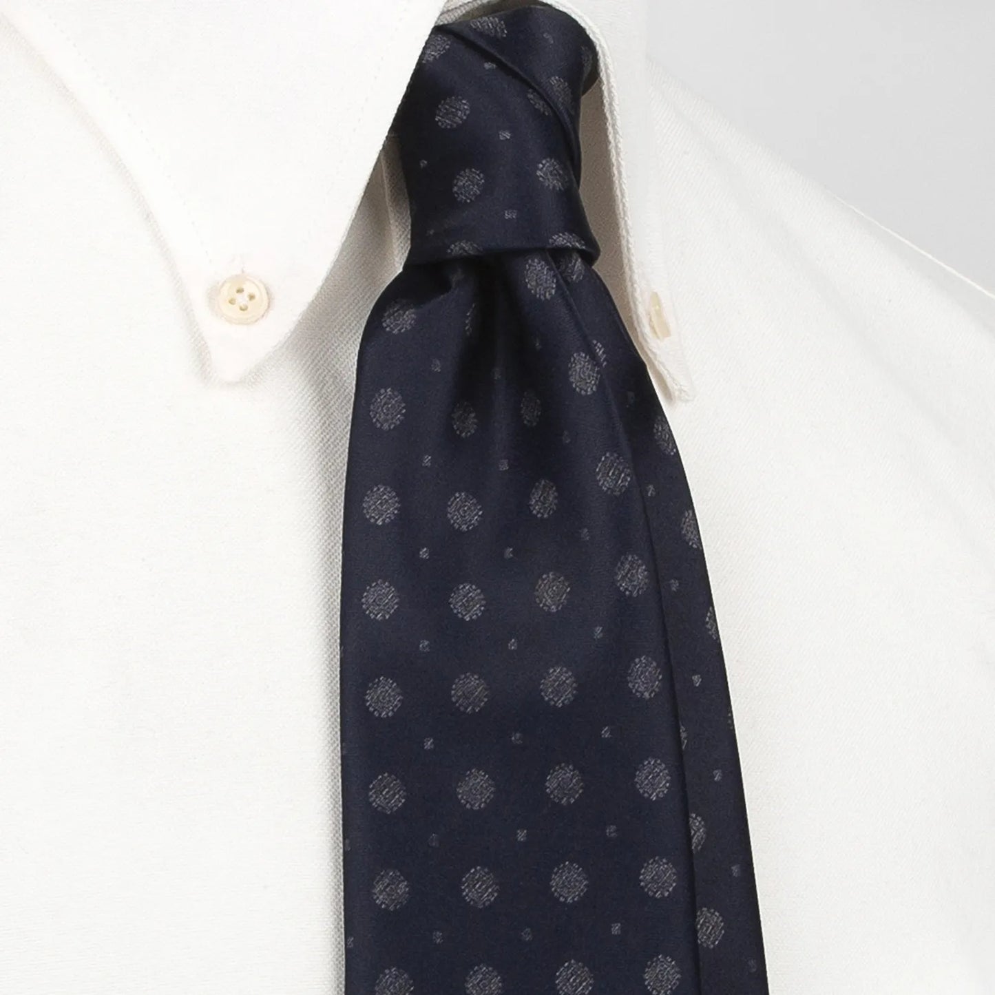 A deep blue men's necktie featuring a subtle pattern of small silver-grey dots and mini floral shapes is tied in a four-in-hand knot on a crisp white button-down shirt, suitable for business and professional settings.