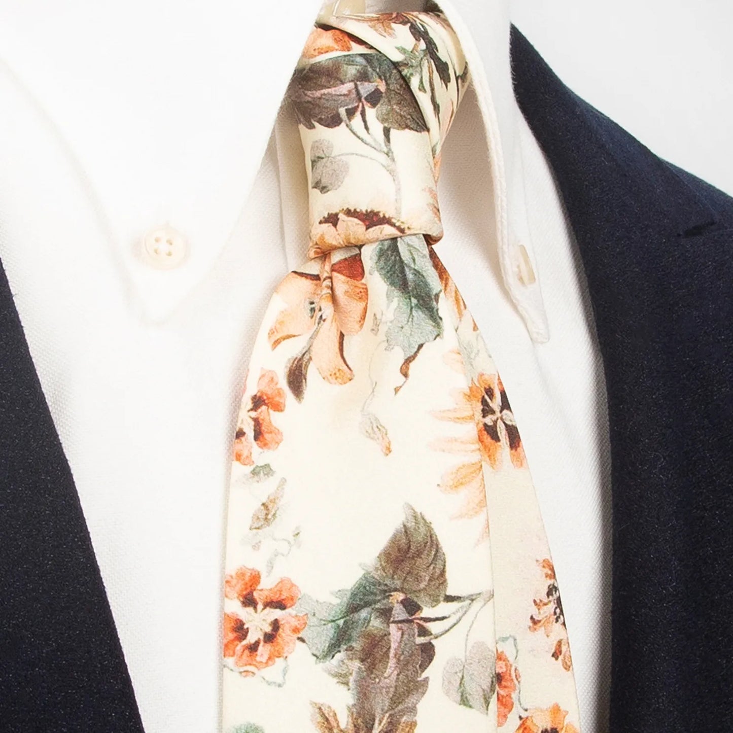 An elegant cream-colored men's necktie from XueDouLing Tie showcasing its classic botanical print with brown, orange, and green flowers and leaves, tied in a four-in-hand knot on a crisp white button-down shirt under a dark blazer, suitable for business casual and stylish events.