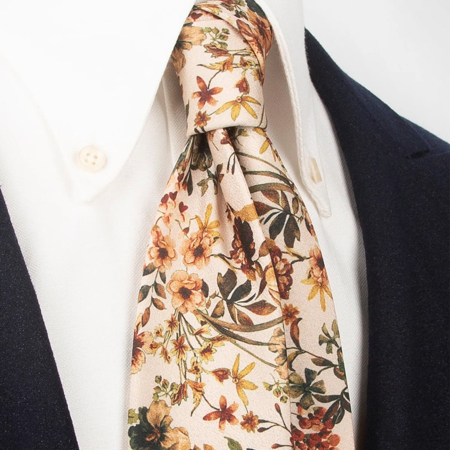 An elegant light beige men's necktie from XueDouLing Tie showcasing its gentle botanical print with brown, yellow, and green flowers and leaves, tied in a four-in-hand knot on a crisp white button-down shirt under a dark blazer, suitable for business casual and graceful events.