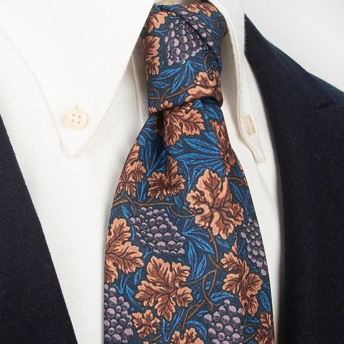 A refined dark blue men's necktie from XueDouLing Tie showcasing its classic grapevine design with brown leaves and light purple grapes, tied in a four-in-hand knot on a crisp white button-down shirt under a dark blazer, suitable for business casual and stylish events.