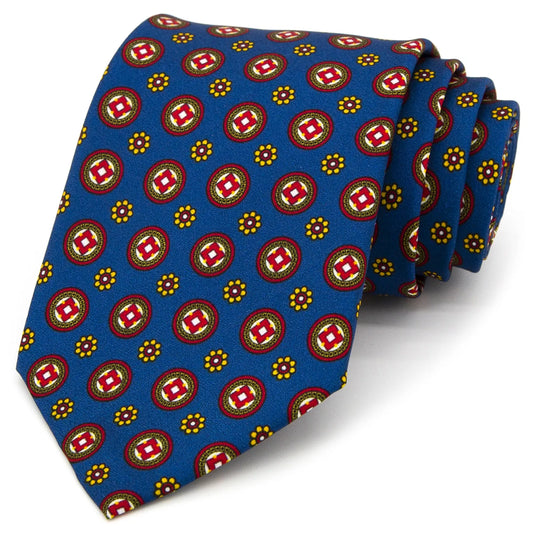 A vibrant blue men's necktie featuring a repeating geometric medallion pattern in red and white, surrounded by small yellow floral dots, is casually knotted against a white background.