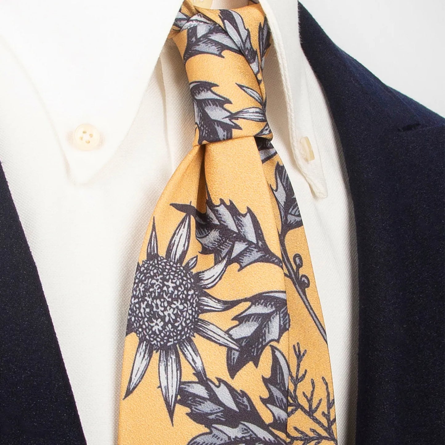 A stylish mustard yellow men's necktie from XueDouLing Tie showcasing its striking botanical design with black and white thistle and cotton motifs, tied in a four-in-hand knot on a crisp white button-down shirt under a dark blazer, suitable for business casual and artistic events.