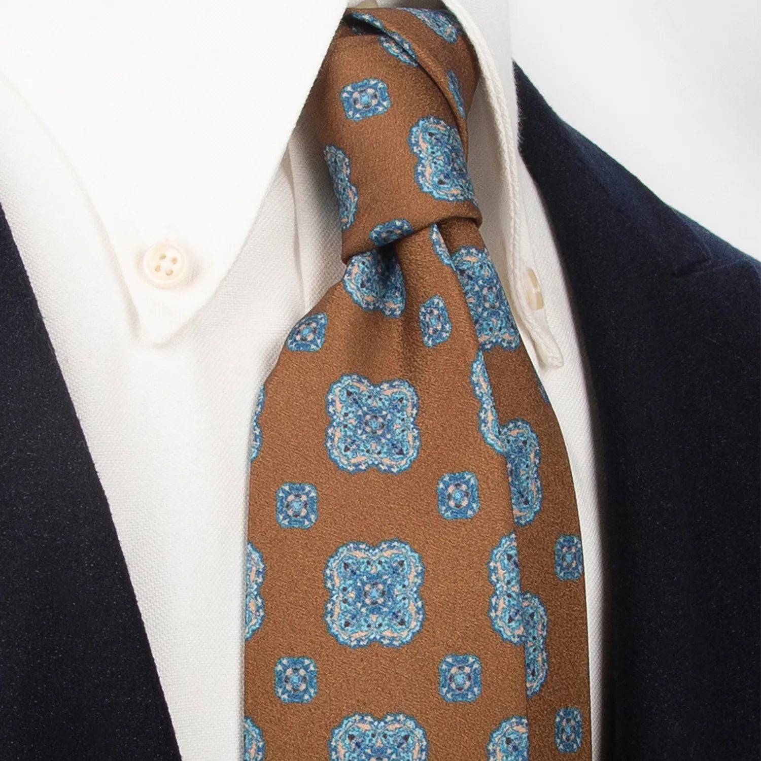 A brown men's necktie with a repeating blue and beige floral motif is tied in a Windsor knot on a white collared shirt under a navy blue blazer.