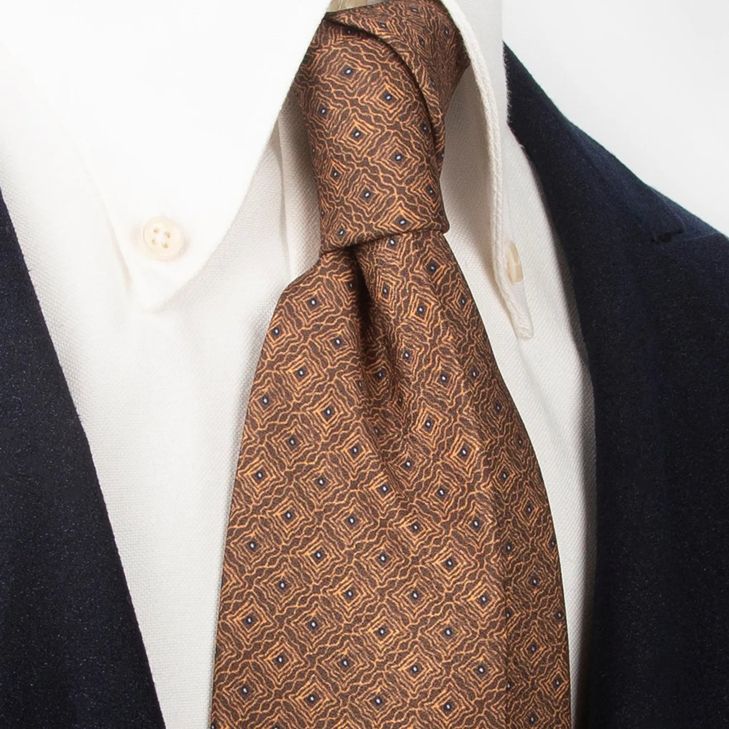 A neatly rolled brown men's necktie with an intricate repeating geometric pattern in gold and dark blue is displayed against a plain white background.
