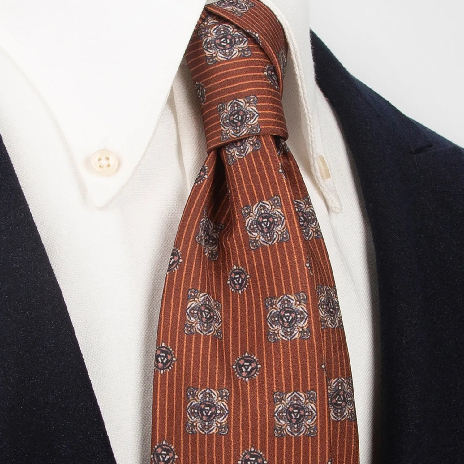 A brown men's necktie with a detailed paisley and stripe pattern is tied in a Windsor knot on a white collared shirt under a dark blue jacket.