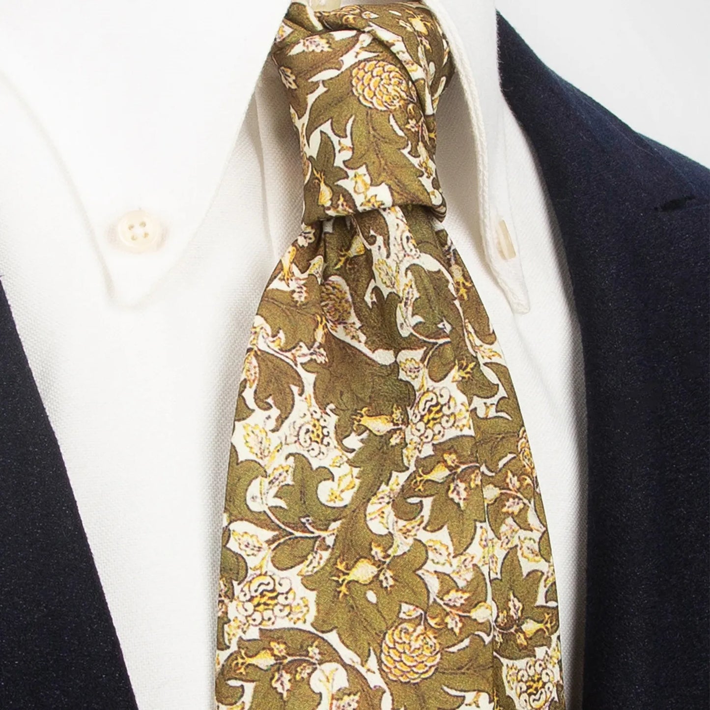 An olive green men's necktie with a rich botanical floral pattern in beige and brown is tied in a four-in-hand knot on a white collared shirt under a dark blue blazer.