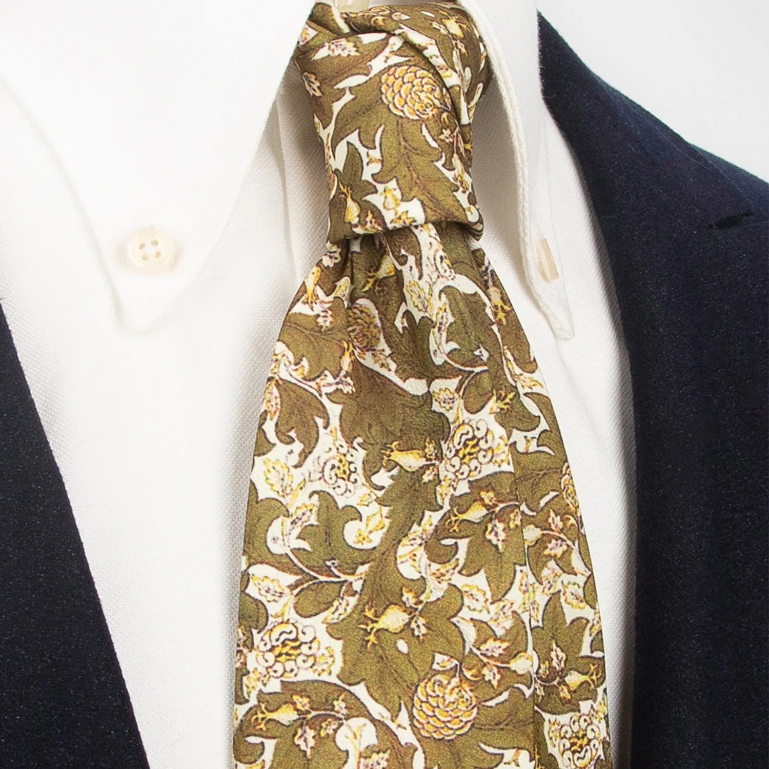 An olive green men's necktie with a rich botanical floral pattern in beige and brown is tied in a four-in-hand knot on a white collared shirt under a dark blue blazer.