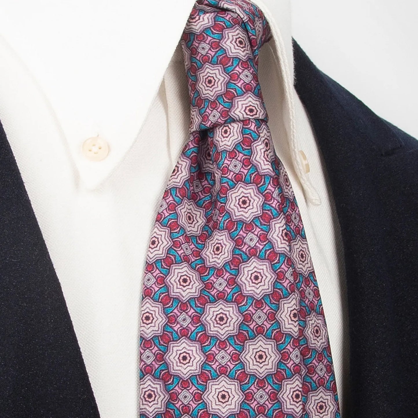 A mauve men's necktie showcasing its intricate geometric pattern in pink, burgundy, and light blue is tied in a Windsor knot on a white collared shirt under a dark blue jacket.