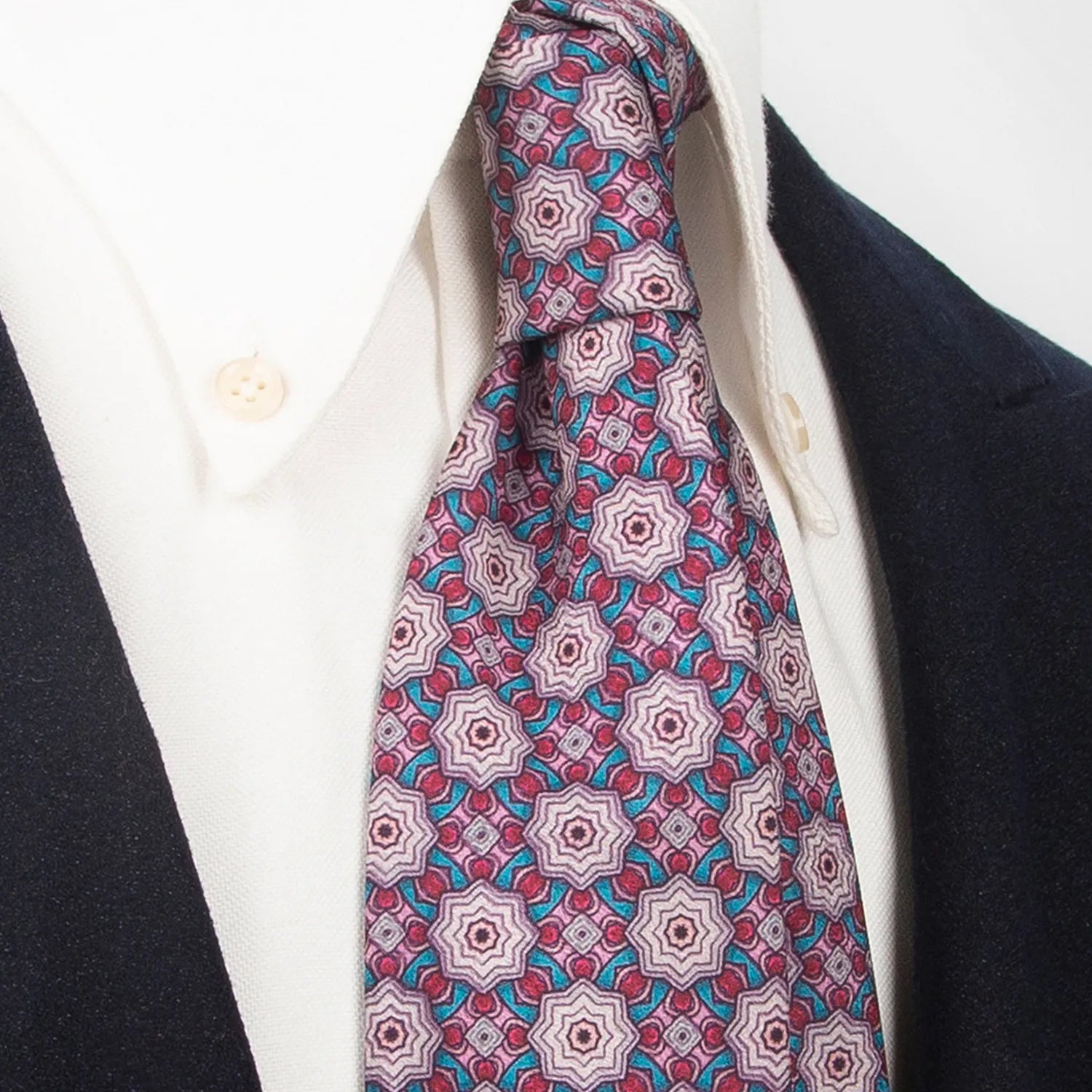 A mauve men's necktie showcasing its intricate geometric pattern in pink, burgundy, and light blue is tied in a Windsor knot on a white collared shirt under a dark blue jacket.