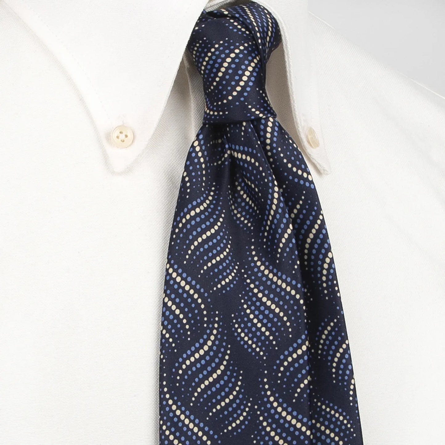 A neatly rolled dark blue men's necktie showcasing its modern wave-like pattern of light blue and beige dots against a clean white backdrop.