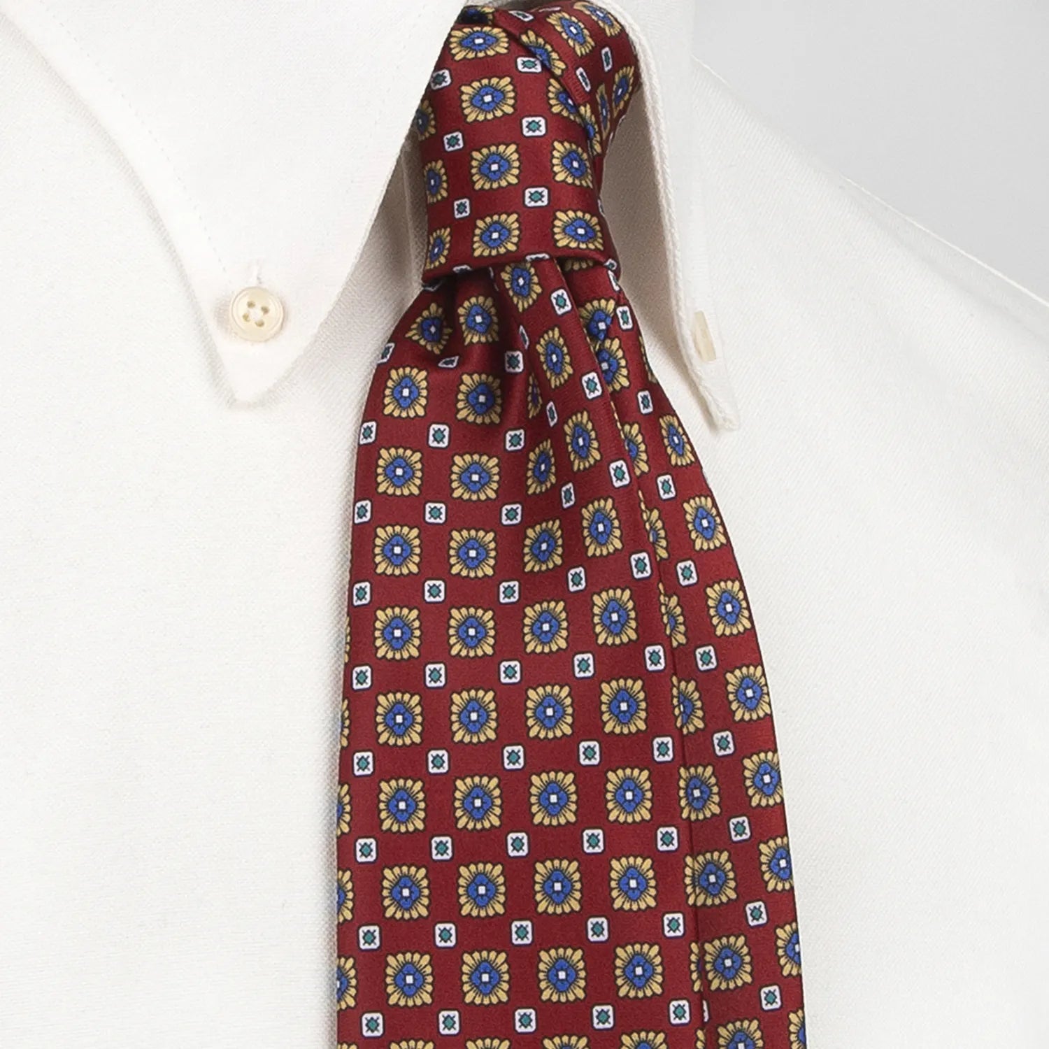 A deep red men's necktie featuring a repeating pattern of small blue and gold flowers with white and green square accents is tied in a Windsor knot on a crisp white button-down shirt.