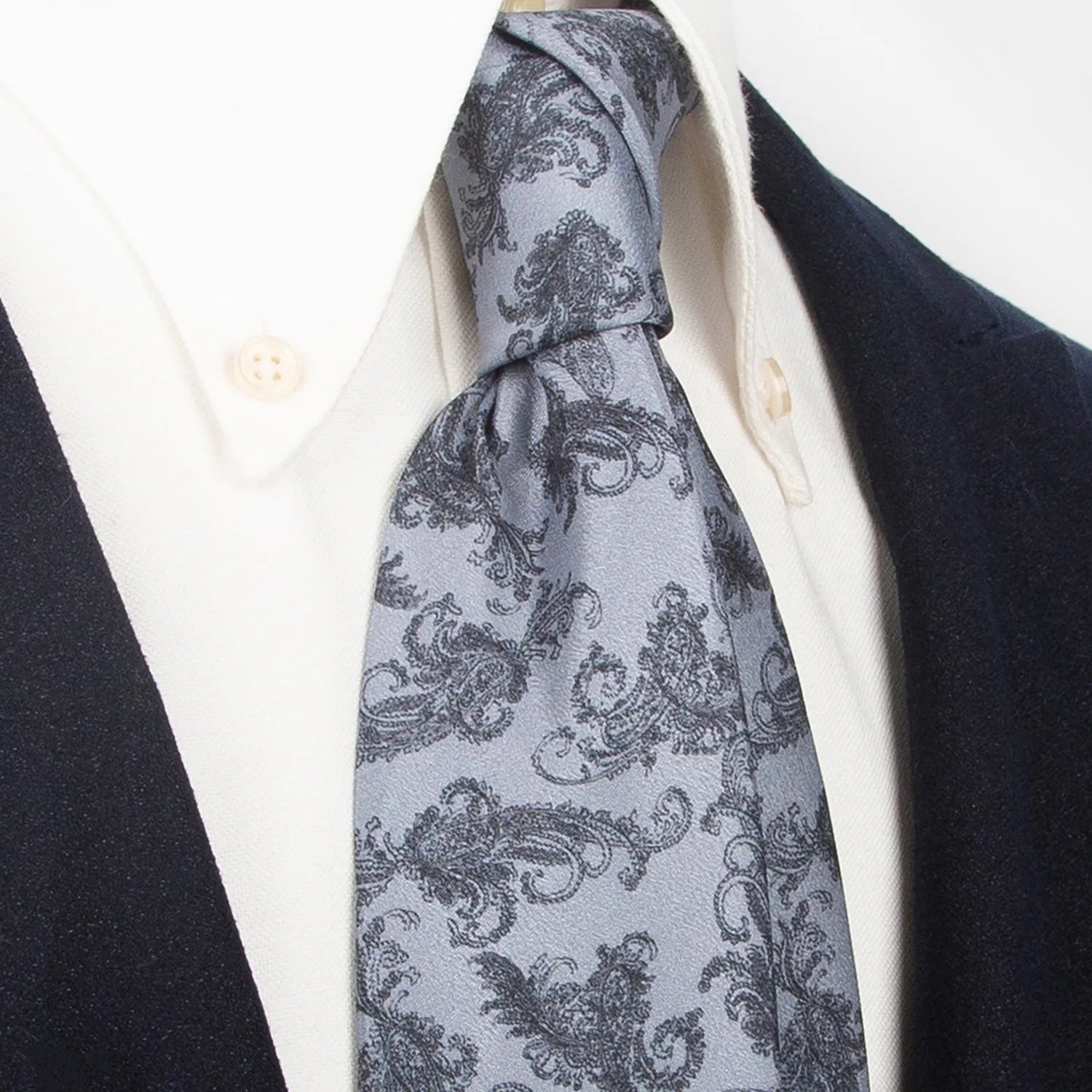 An elegant light grey men's necktie from XueDouLing Tie with a classic black paisley design is tied in a Windsor knot on a crisp white button-down shirt under a dark navy blazer, suitable for business and events.