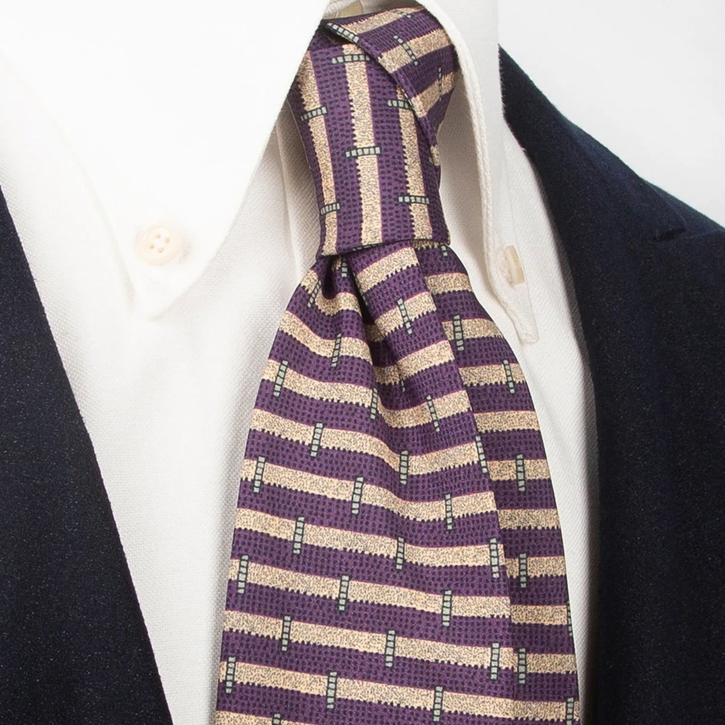 A stylish deep purple men's necktie featuring a unique geometric bar stripe design in beige and black is tied in a four-in-hand knot on a crisp white button-down shirt under a dark navy blazer, suitable for business and events.