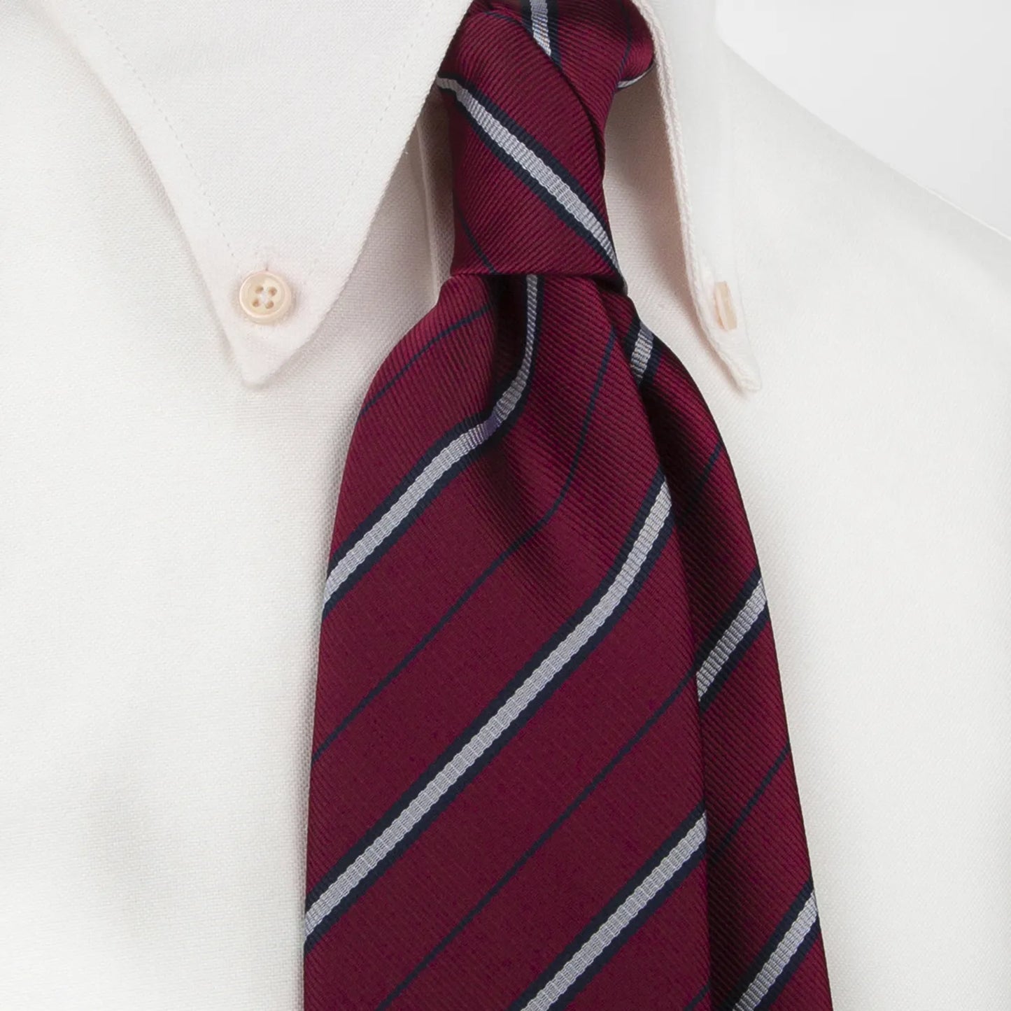 A burgundy men's necktie featuring bold diagonal stripes in silver and navy blue is tied in a four-in-hand knot on a crisp white button-down shirt, suitable for business and meetings.