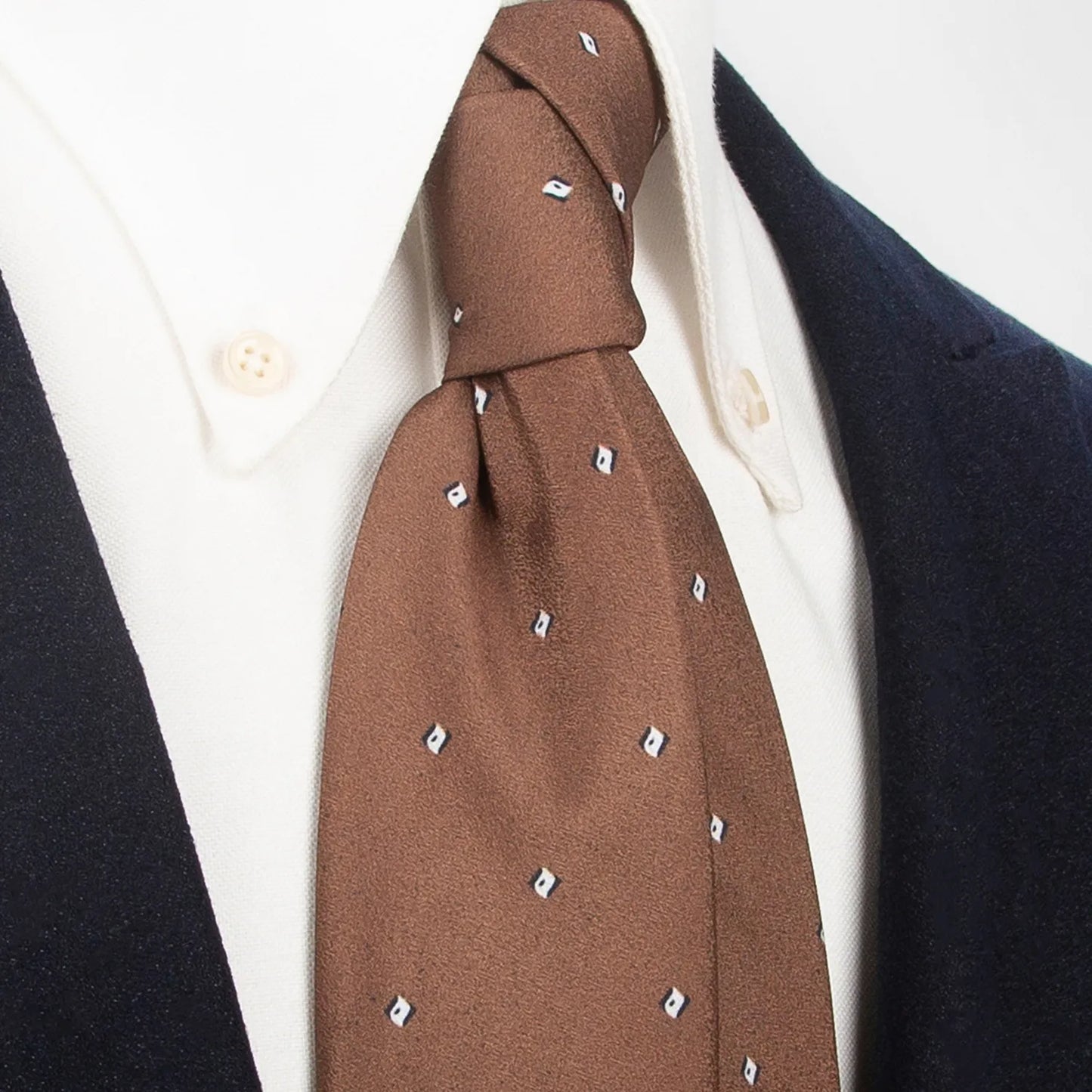 A refined brown men's necktie from XueDouLing Tie with a minimalist pattern of small white and blue geometric shapes is tied in a four-in-hand knot on a crisp white button-down shirt under a dark suit jacket, suitable for business and understated elegance.