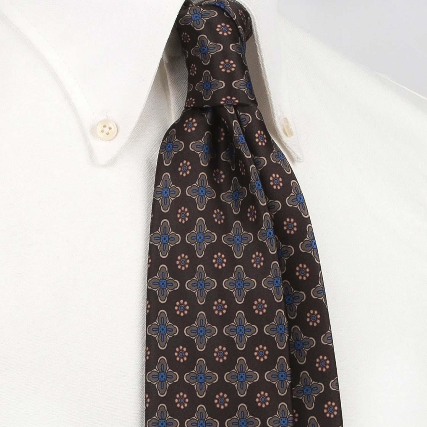 A neatly rolled dark brown men's necktie showcasing its subtle yet intricate pattern of small blue, beige, and brown floral and geometric designs against a clean white backdrop