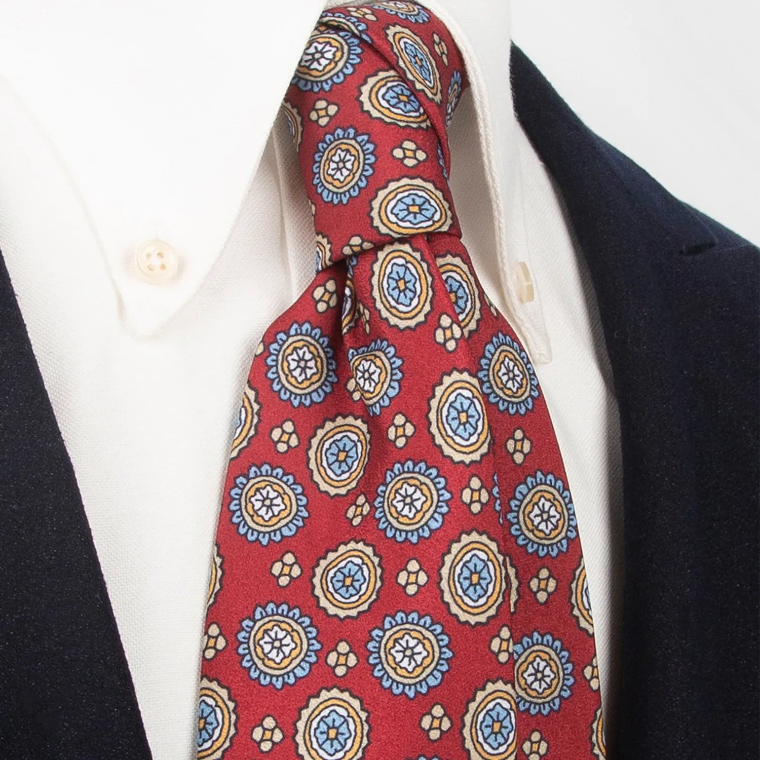 An elegant deep red men's necktie from XueDouLing Tie with a lively pattern of circular floral medallions in blue, yellow, and white, tied in a four-in-hand knot on a crisp white button-down shirt under a dark suit jacket, perfect for business, weddings, and festive events.