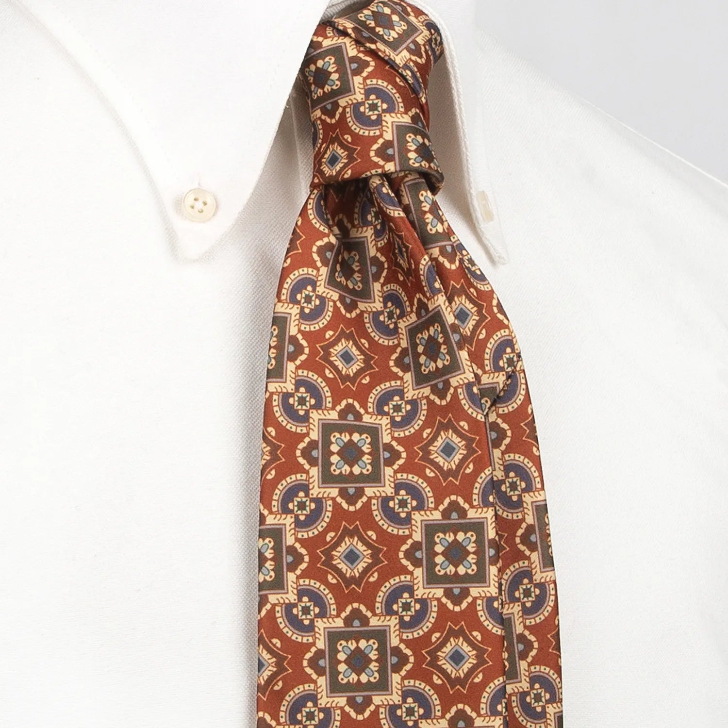 A red-brown men's necktie featuring a complex geometric pattern in blue, beige, and green, incorporating squares, circles, and star-like shapes, is tied in a four-in-hand knot on a crisp white button-down shirt, suitable for business and elegant events.