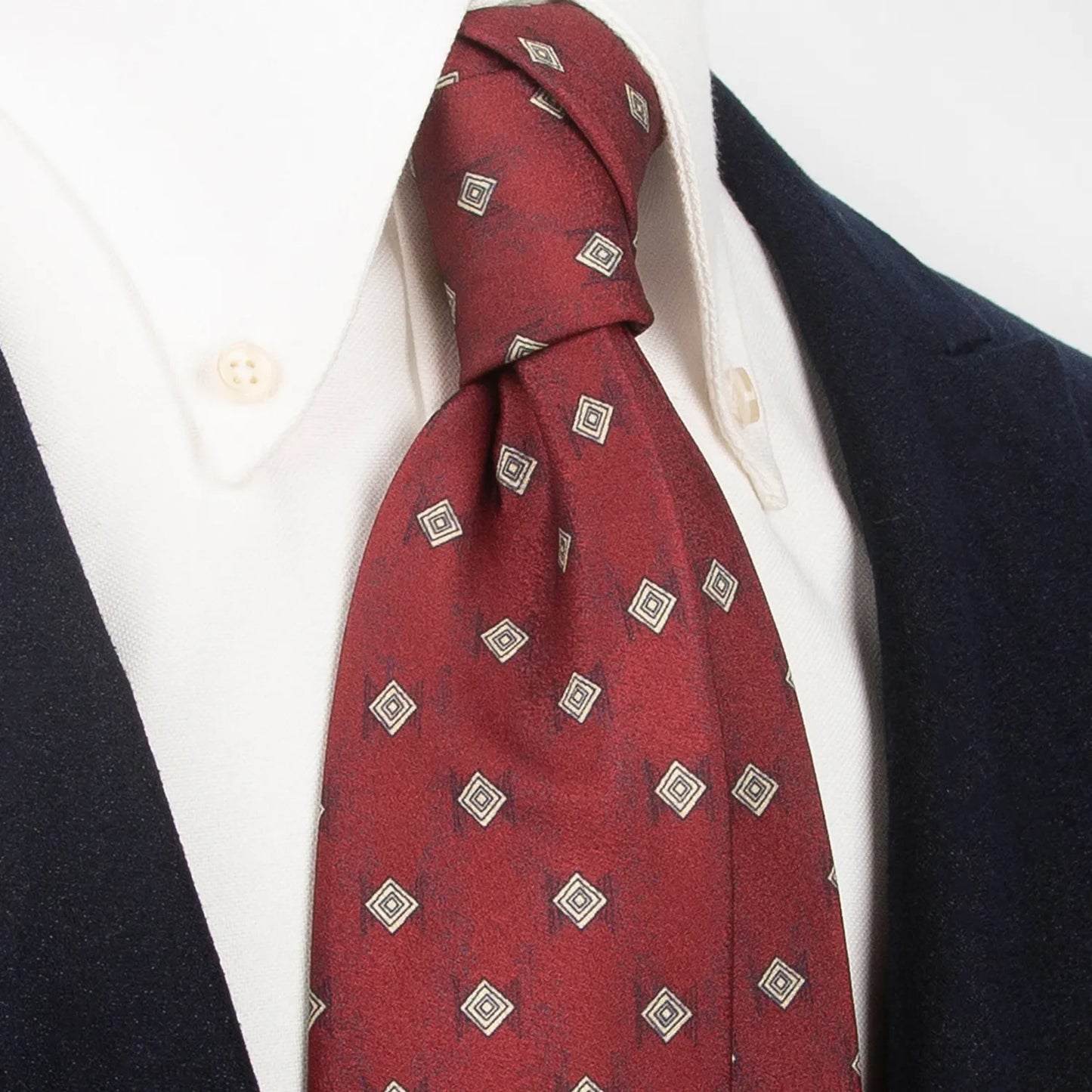 An elegant deep red men's necktie from XueDouLing Tie with a subtle geometric diamond design in beige and black is tied in a four-in-hand knot on a crisp white button-down shirt under a dark blazer, suitable for business and refined events.