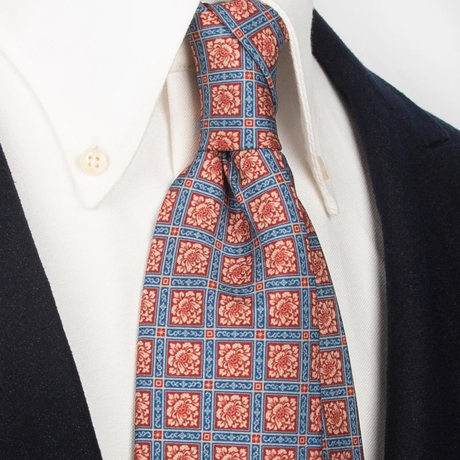 A neatly folded red-brown, blue, and beige men's necktie showcasing its complex geometric floral pattern against a clean white surface.