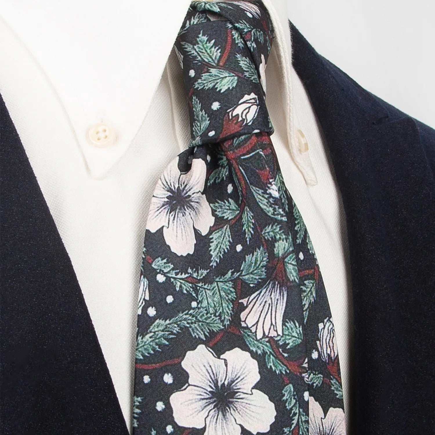 A deep black men's necktie featuring a minimalist floral pattern with white blossoms and green leaves, accented by small white dots, is casually knotted against a white background.