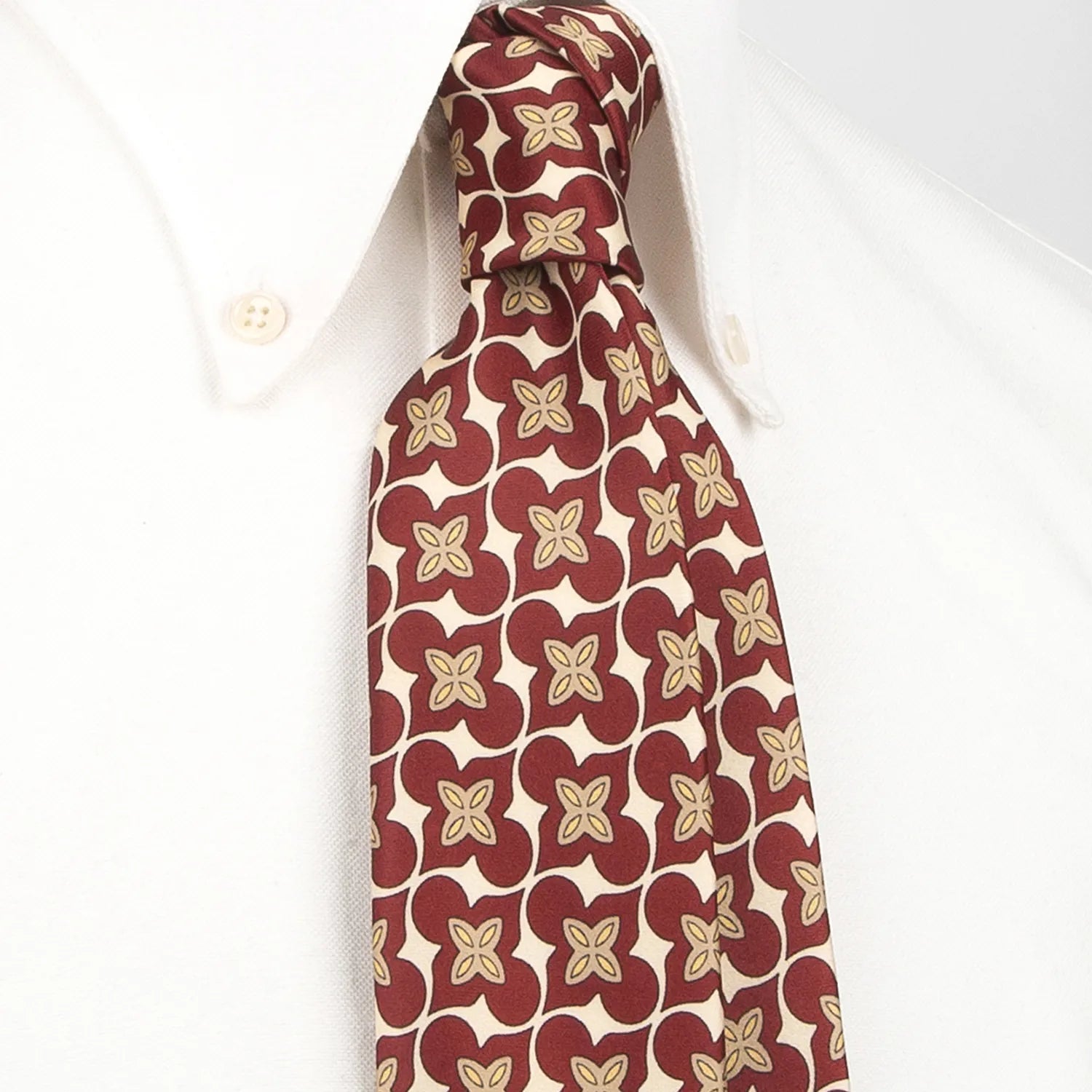 A deep red men's necktie featuring an elegant floral and curved geometric pattern in beige and gold is tied in a four-in-hand knot on a crisp white button-down shirt, suitable for business and refined events.