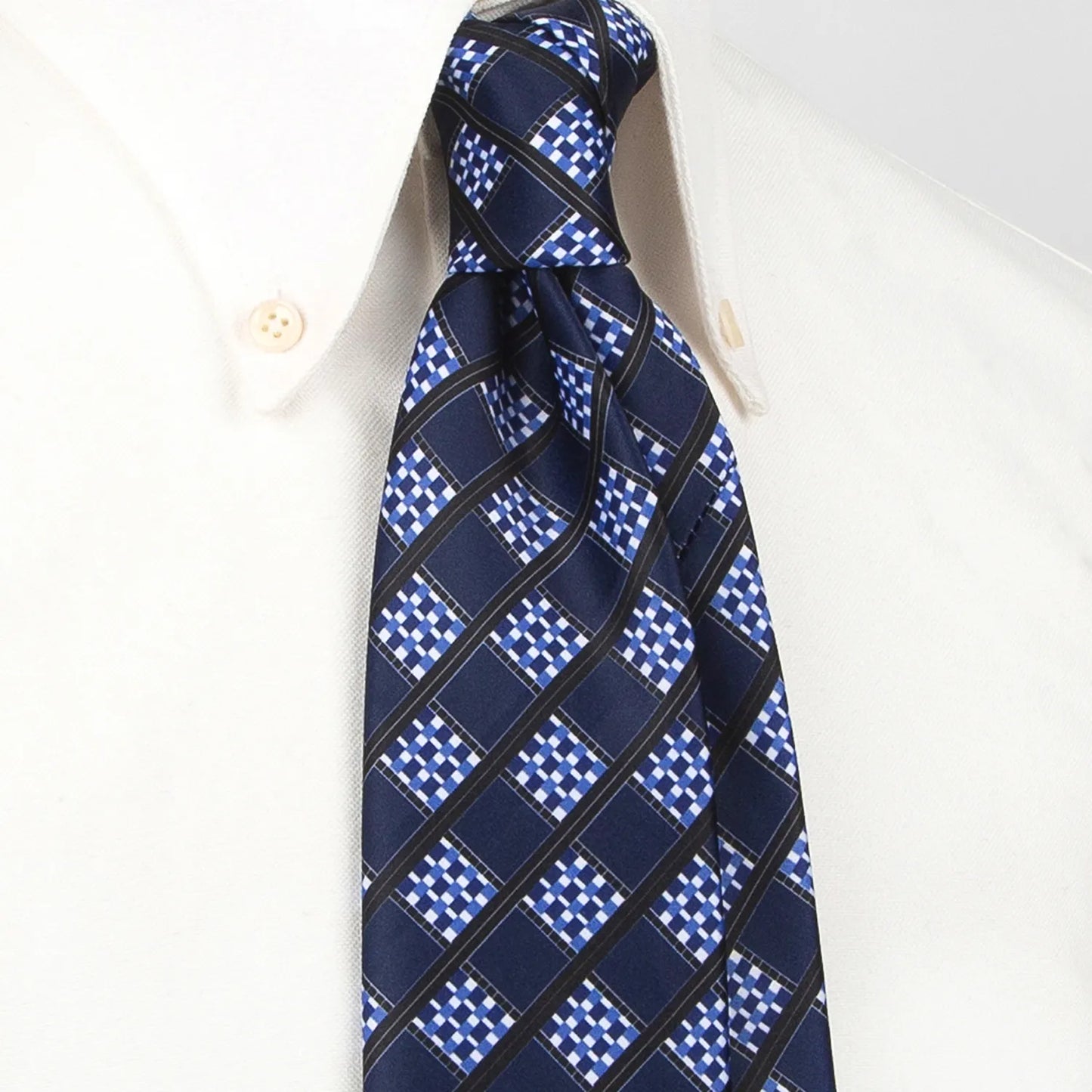 A deep blue men's necktie featuring a modern angled grid pattern in white, light blue, and black is tied in a four-in-hand knot on a crisp white button-down shirt, suitable for business and contemporary style.