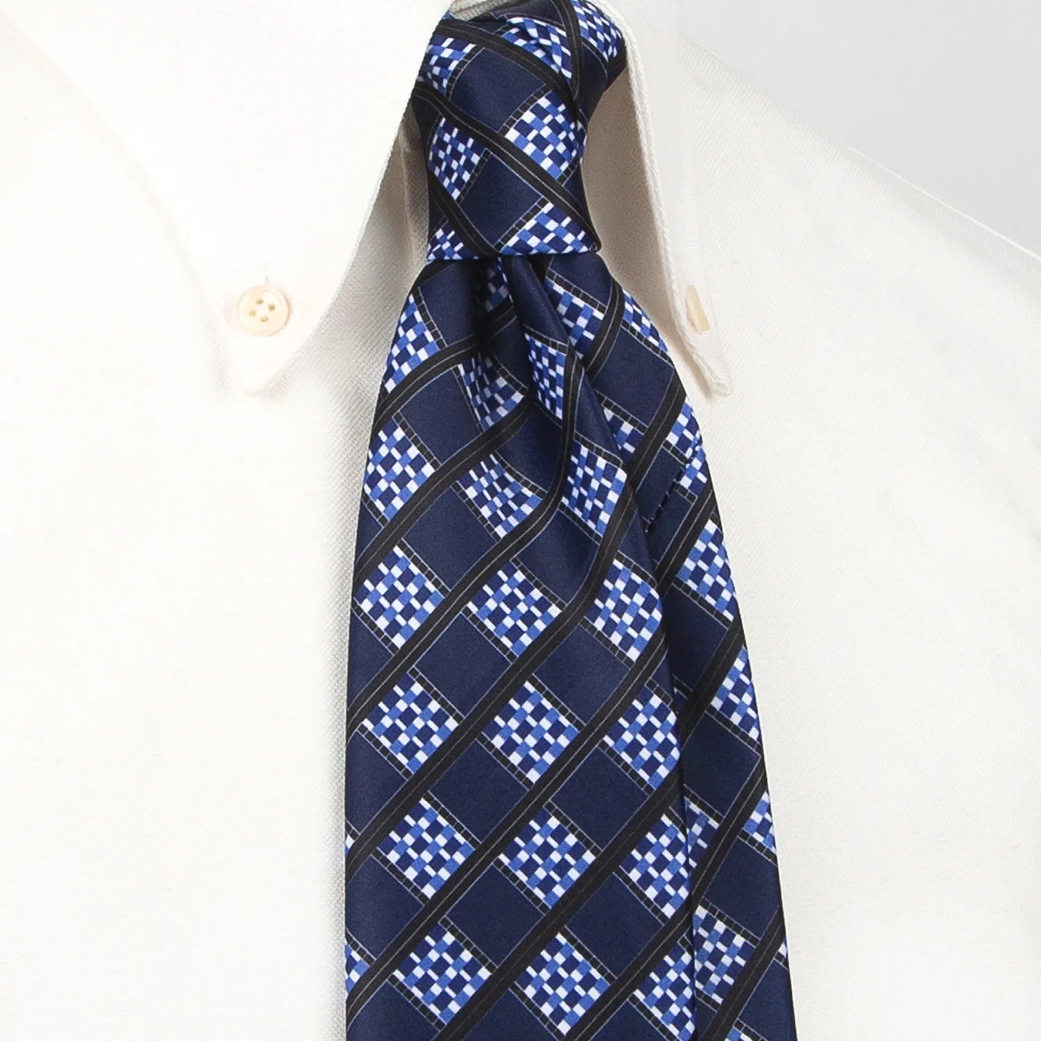 A deep blue men's necktie featuring a modern angled grid pattern in white, light blue, and black is tied in a four-in-hand knot on a crisp white button-down shirt, suitable for business and contemporary style.