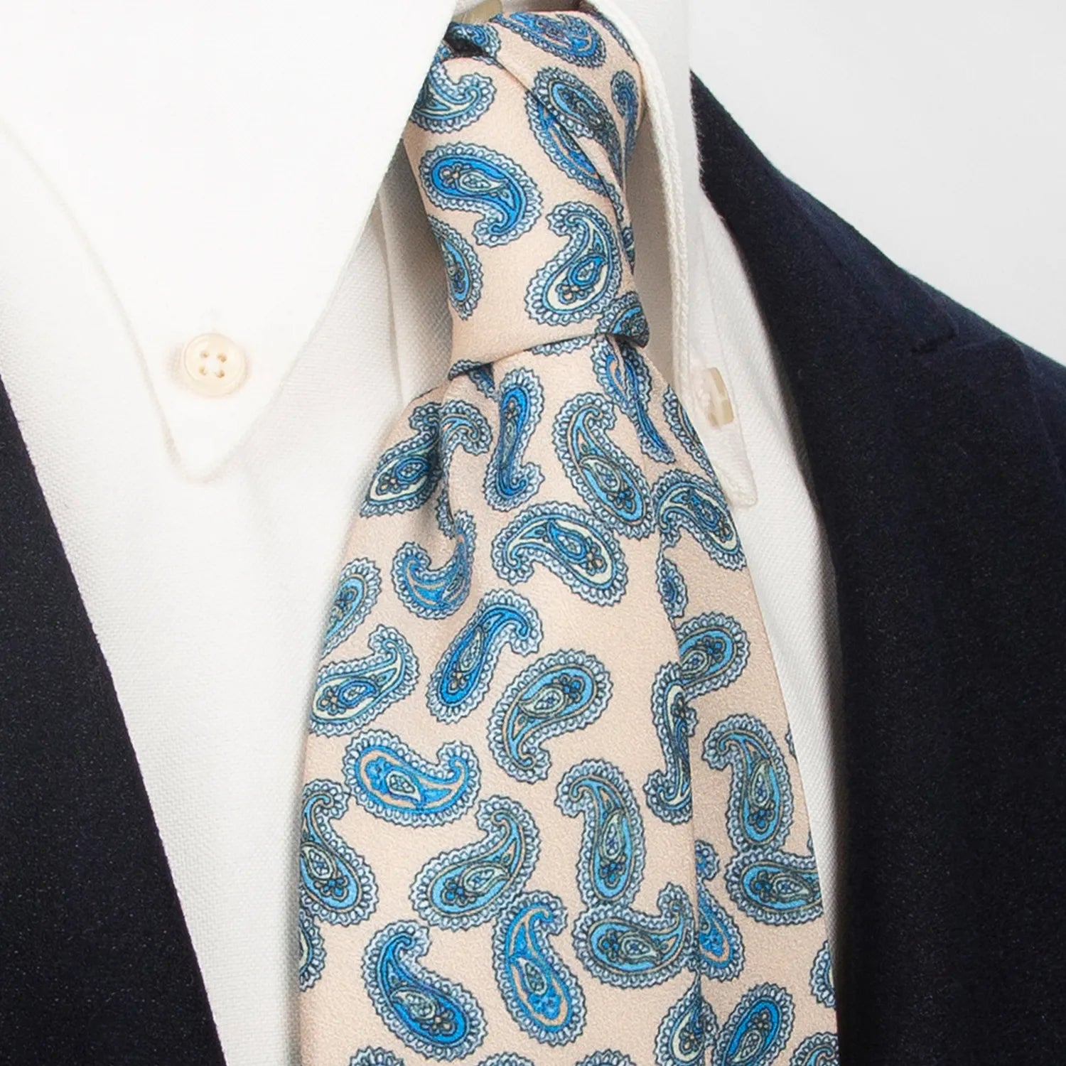 A refined beige men's necktie from XueDouLing Tie showcasing its elegant blue paisley design, accented with yellow, tied in a four-in-hand knot on a crisp white button-down shirt under a dark blazer, suitable for business casual and stylish events.