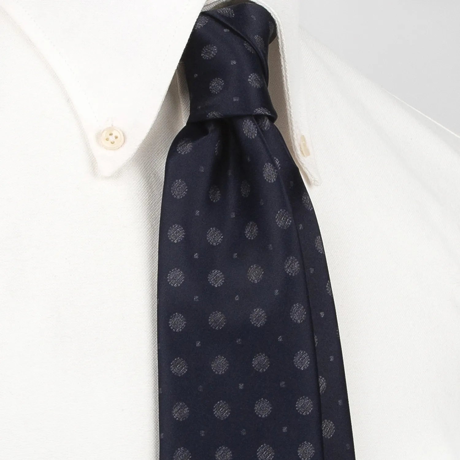 A deep blue men's necktie featuring a subtle pattern of small silver-grey dots and mini floral shapes is tied in a four-in-hand knot on a crisp white button-down shirt, suitable for business and professional settings.