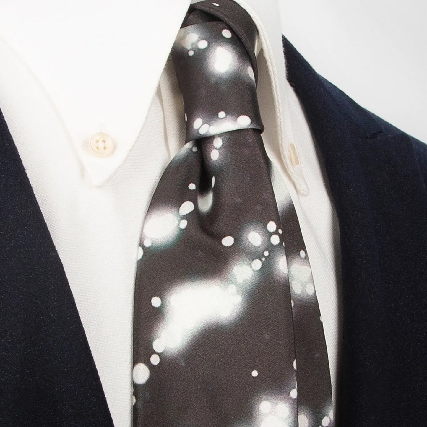 A unique dark brown men's necktie from XueDouLing Tie showcasing its abstract white and light green ink blot design, tied in a four-in-hand knot on a crisp white button-down shirt under a dark blazer, suitable for business casual and artistic events.