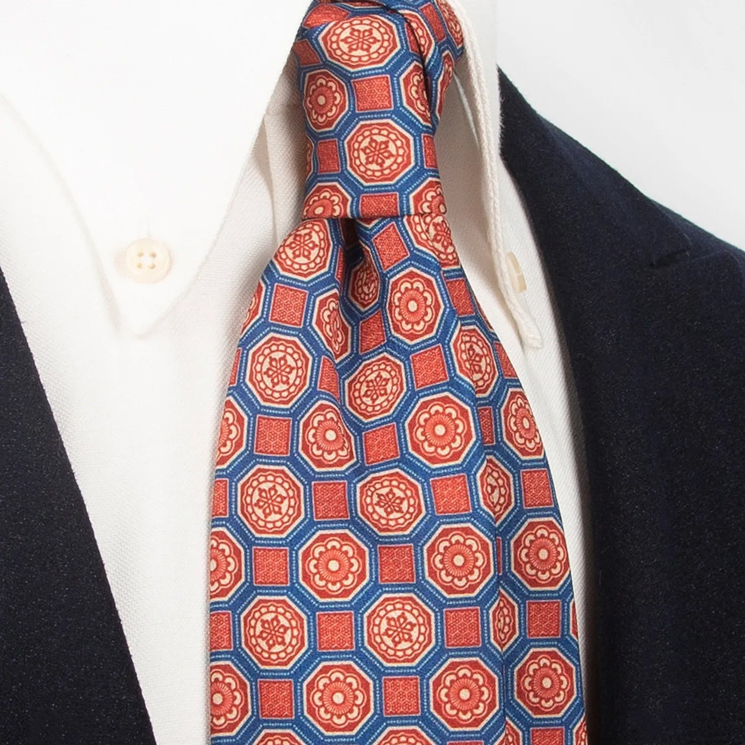 A blue men's necktie featuring an intricate geometric floral pattern in red, beige, and brown is tied in a four-in-hand knot on a crisp white button-down shirt, suitable for business and stylish events.