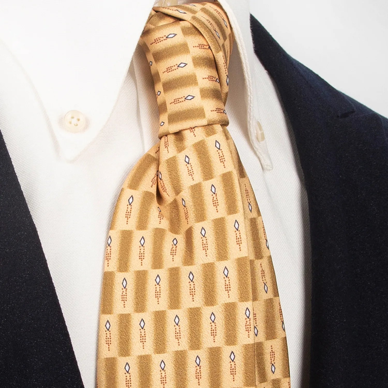 A gold and brown checkered men's necktie featuring an abstract geometric pattern in white, blue, and brown is tied in a four-in-hand knot on a crisp white button-down shirt, suitable for business and modern style.