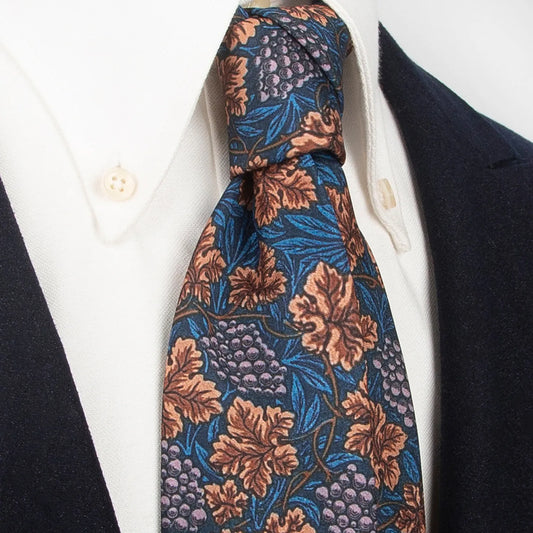 A refined dark blue men's necktie from XueDouLing Tie showcasing its classic grapevine design with brown leaves and light purple grapes, tied in a four-in-hand knot on a crisp white button-down shirt under a dark blazer, suitable for business casual and stylish events.